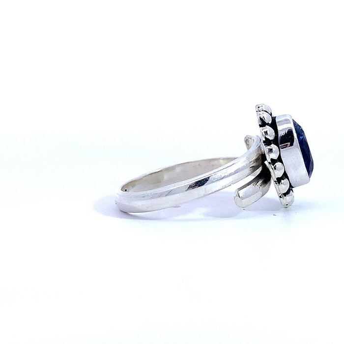 photo of a silver ring set with an oval blue gem finished with a beaded halo on a white background side view