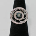 Silver ring with a central pearl and diamond-like stones on a black stand against a gray background