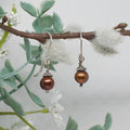 Pair of brown pearl earrings on a branch with green leaves and white flowers.