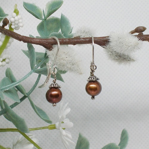 Pair of brown pearl earrings on a branch with green leaves and white flowers.