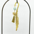 Gold feather-shaped metal bookmark with a green bead and gold tassel on a white background