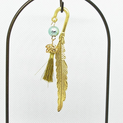 Gold feather-shaped metal bookmark with a green bead and gold tassel on a white background