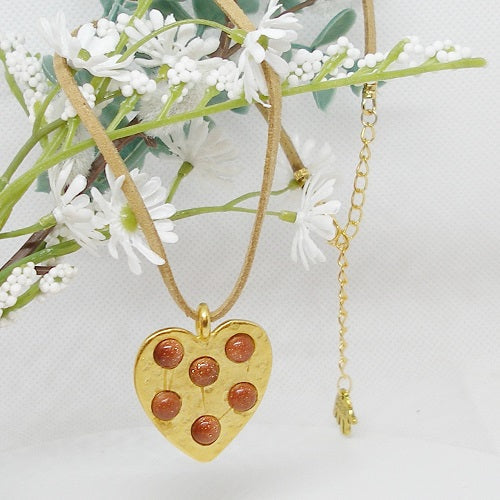 Gold heart-shaped pendant with embedded stones on a gold chain, displayed against a white background with greenery.
