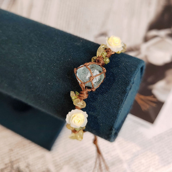 The BohoBud Bracelet brings the softest parts of nature to your wrist. Featuring a glacier-blue synthetic crystal as the centerpiece, wrapped in rustic brown thread, this bracelet is adorned with tiny gemstone chips and mini white rose buds that look freshly picked from a meadow. Each knot is intentional, each detail handwoven, making it feel like a warm afternoon hug.