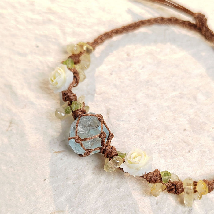 The BohoBud Bracelet brings the softest parts of nature to your wrist. Featuring a glacier-blue synthetic crystal as the centerpiece, wrapped in rustic brown thread, this bracelet is adorned with tiny gemstone chips and mini white rose buds that look freshly picked from a meadow. Each knot is intentional, each detail handwoven, making it feel like a warm afternoon hug.