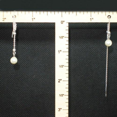 Pearl earrings with chains hanging on a ruler for size reference against a black background