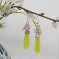 Green teardrop earrings with pink beads hanging from a branch with white flowers.