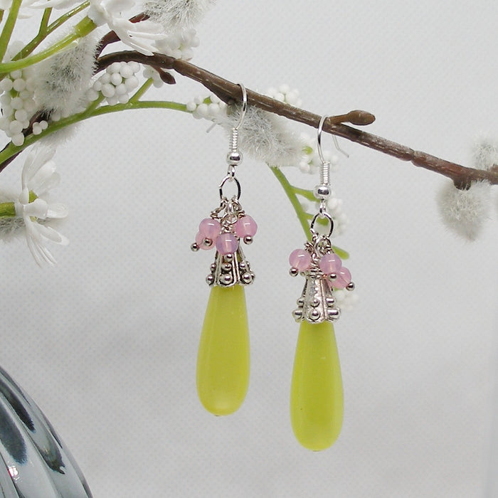 Green teardrop earrings with pink beads hanging from a branch with white flowers.