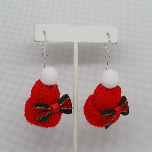 Red knitted earrings with white pom-poms and red and green bows on a white stand against a light gray background.