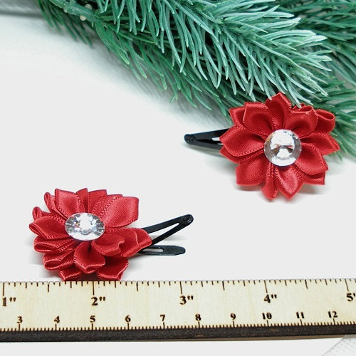 Two red flower hair clips with a central gemstone on a white surface with greenery in the background.