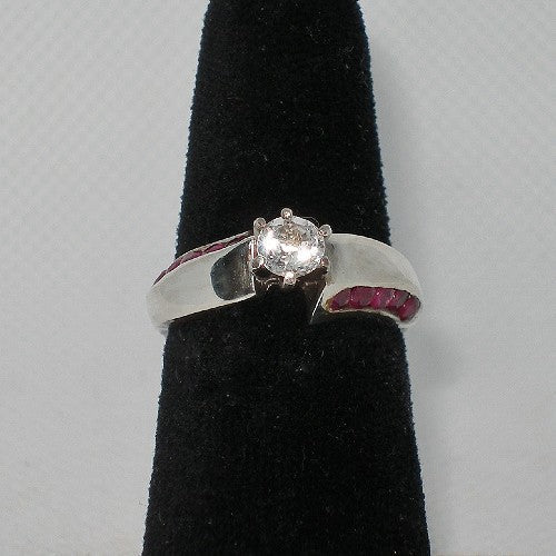 Silver ring with a white gem and ruby stones on a black stand against a gray background