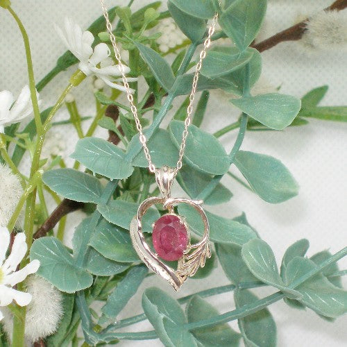 Silver pendant with a red gemstone on a chain, set against green leaves and white flowers.