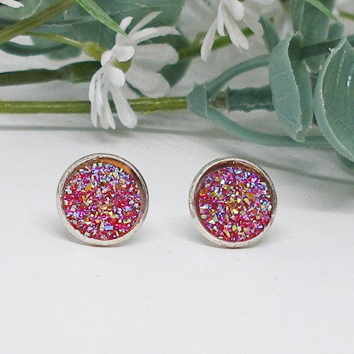 Pair of round stud earrings with red glittery centers on a white surface with green leaves in the background.