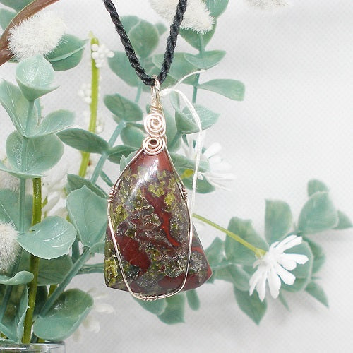 Necklace with a pendant featuring red and green colors, hanging against a natural background with green leaves and white flowers.