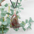 Necklace with a pendant featuring red and green colors, hanging against a natural background with green leaves and white flowers.
