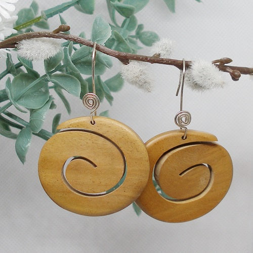 Wooden spiral earrings hanging on a branch with green leaves and white flowers.